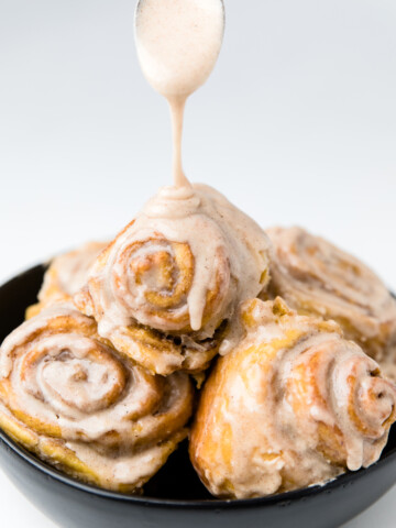 a black bowl of dairy free pumpkin cinnamon rolls with glaze drizzling from the top.