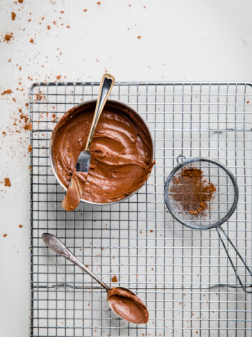 a bowl of dairy free chocolate frosting and a sieve of cocoa powder on a rack.