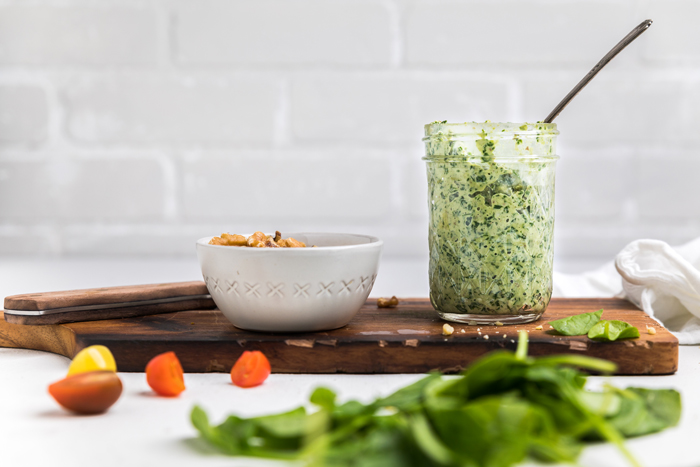 a jar of dairy free pesto on a wooden board next to a white bowl of walnuts.