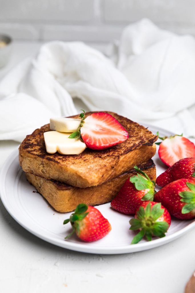 two slices of dairy free french toast stacked on a plate with butter and fresh strawberries.