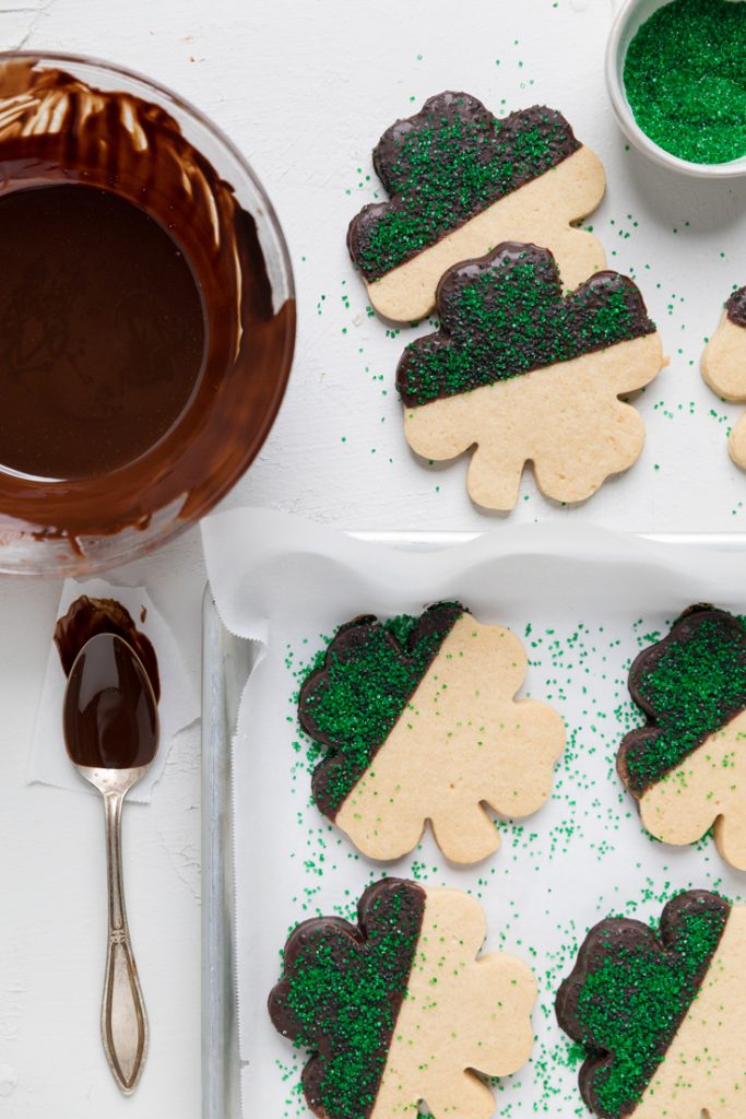 shamrock shaped vegan sugar cookies dipped in chocolate with green sugar crystals on half.