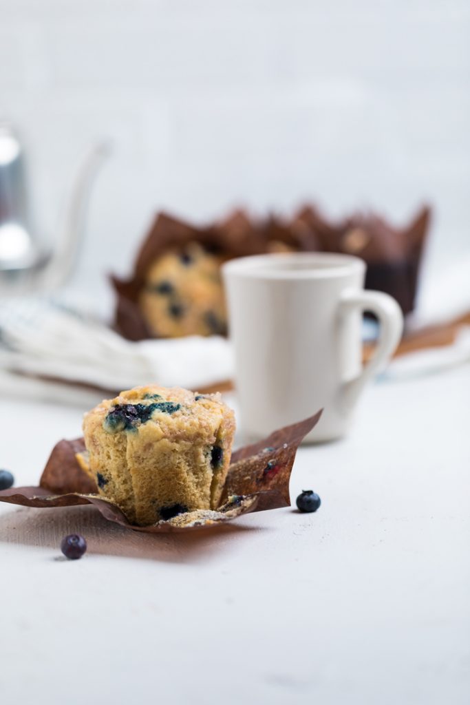 a single vegan blueberry muffin on a wrapper with a white mug behind it.