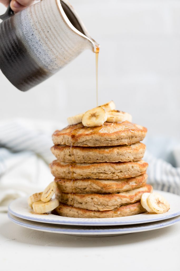 A stack of fluffy Vegan banana pancakes topped with fresh banana slices with syrup drizzling overtop.