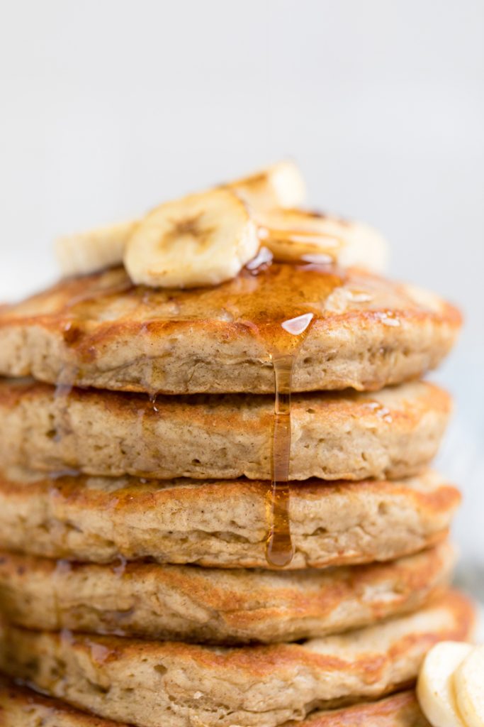 four fluffy Vegan banana pancakes topped with fresh banana slices with syrup drizzling down.