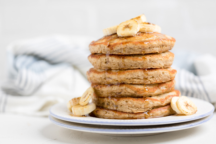 six Vegan banana pancakes topped with fresh banana slices with syrup drizzling overtop.