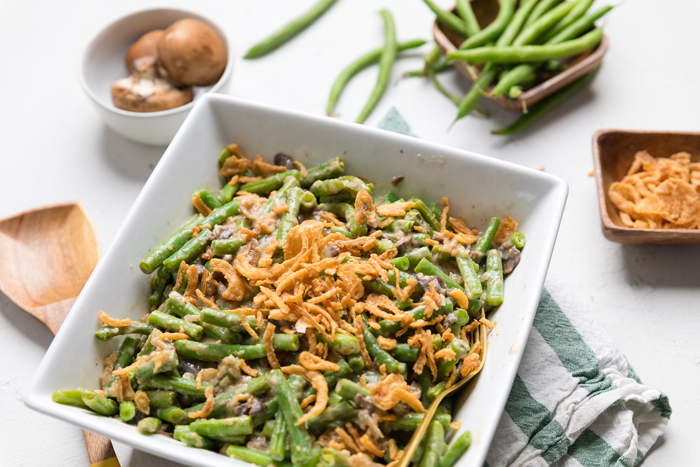Vegan green bean casserole in a dish on a table with bowls of mushrooms and uncooked green beans on the side.