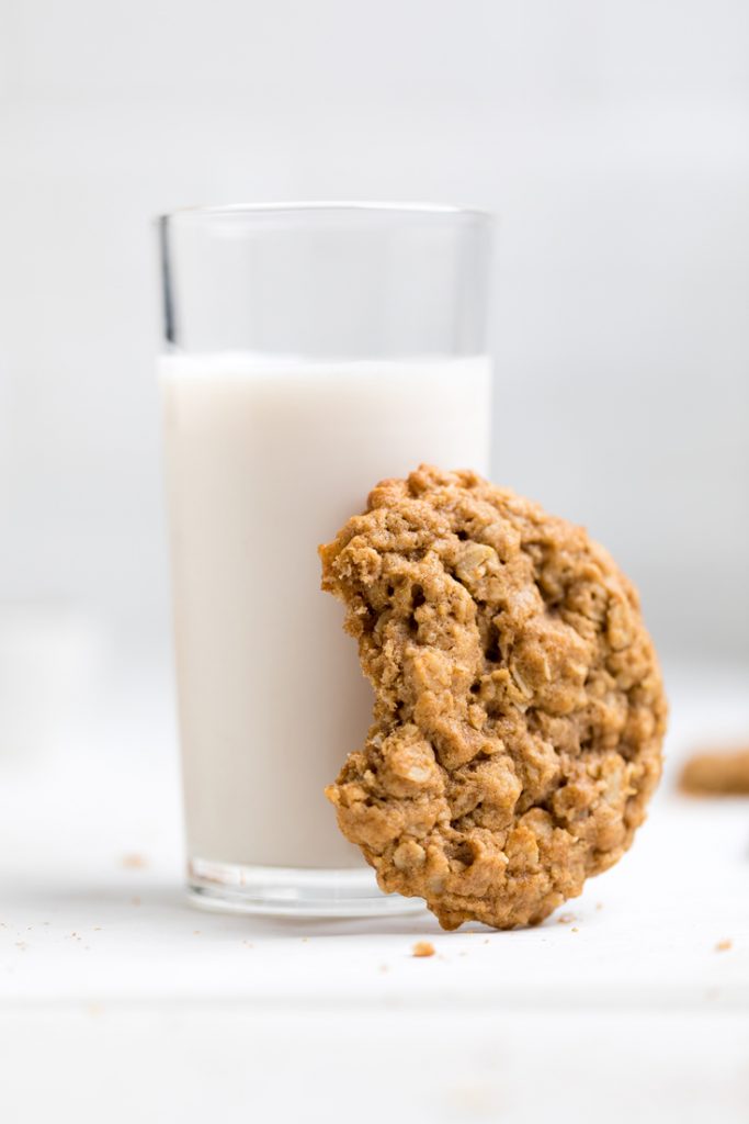 a vegan oatmeal cookie with a bite out resting against a glass of non-dairy milk.