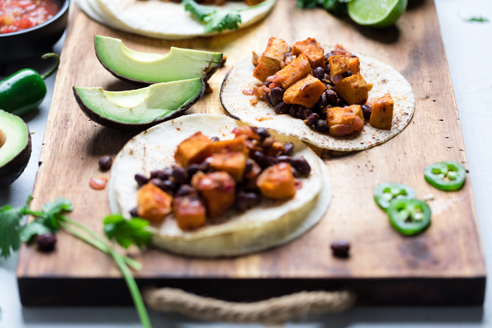 two vegan sweet potato and black bean tacos on a wooden board with avocado wedges.
