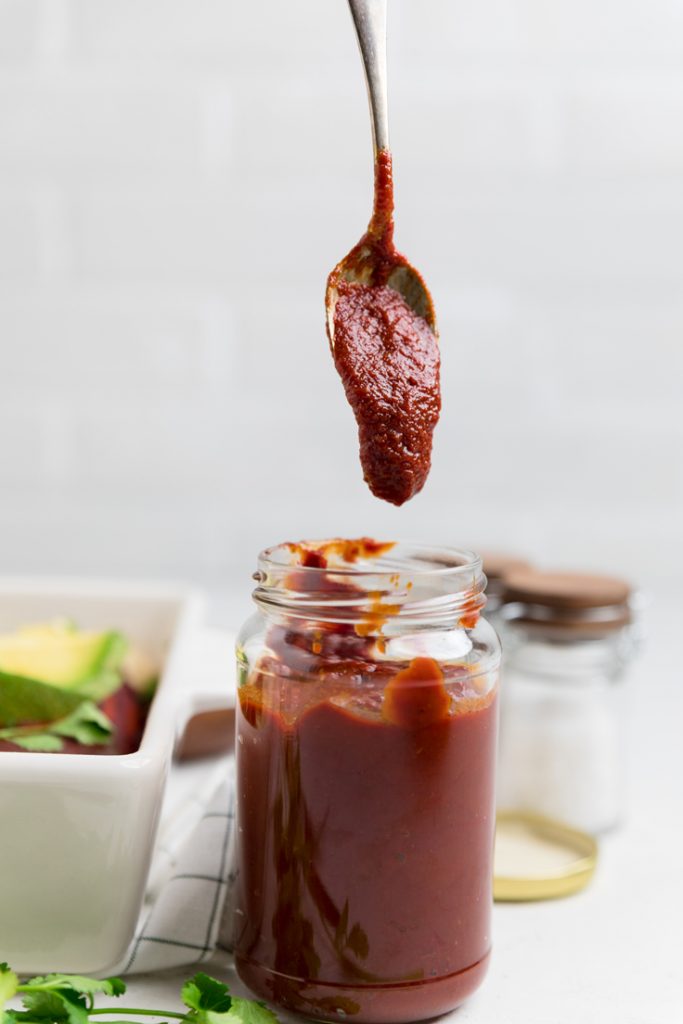 a spoon of red enchilada sauce hovering over a glass jar.