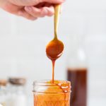 a hand holding a spoon of homemade vegan bbq sauce over a jar.