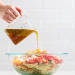 A hand pouring dressing from a measuring cup over vegan pasta salad ingredients.