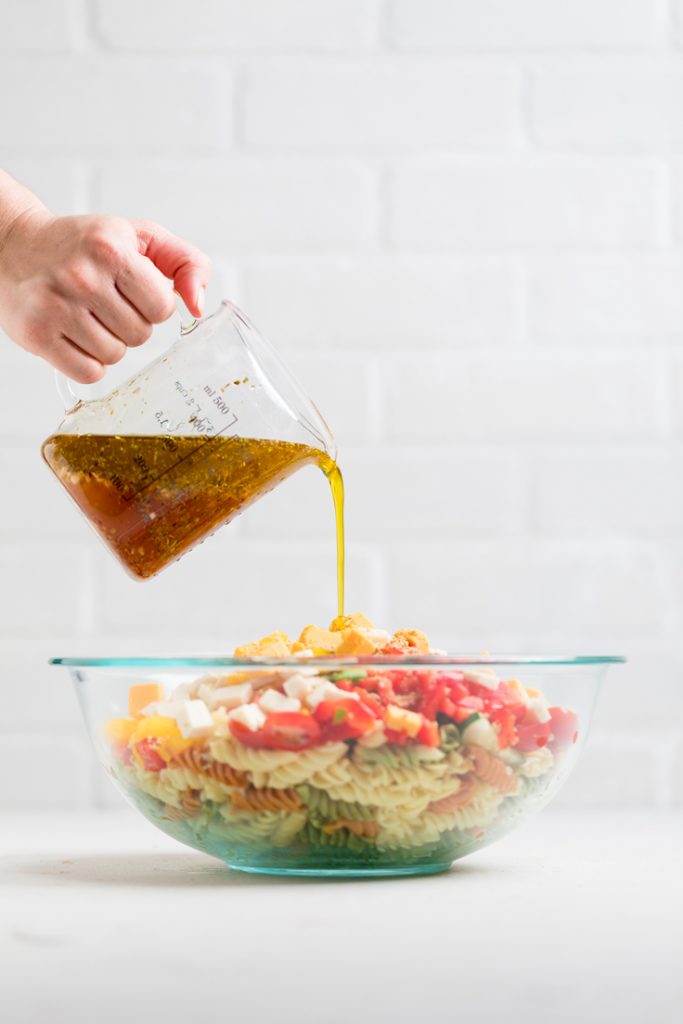 A hand pouring dressing from a measuring cup over vegan pasta salad ingredients.