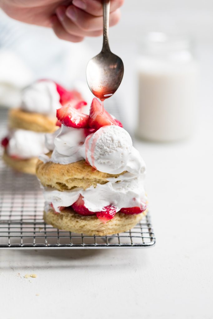 a vegan strawberry shortcake on a wire rack with strawberry syrup being drizzled from a spoon.