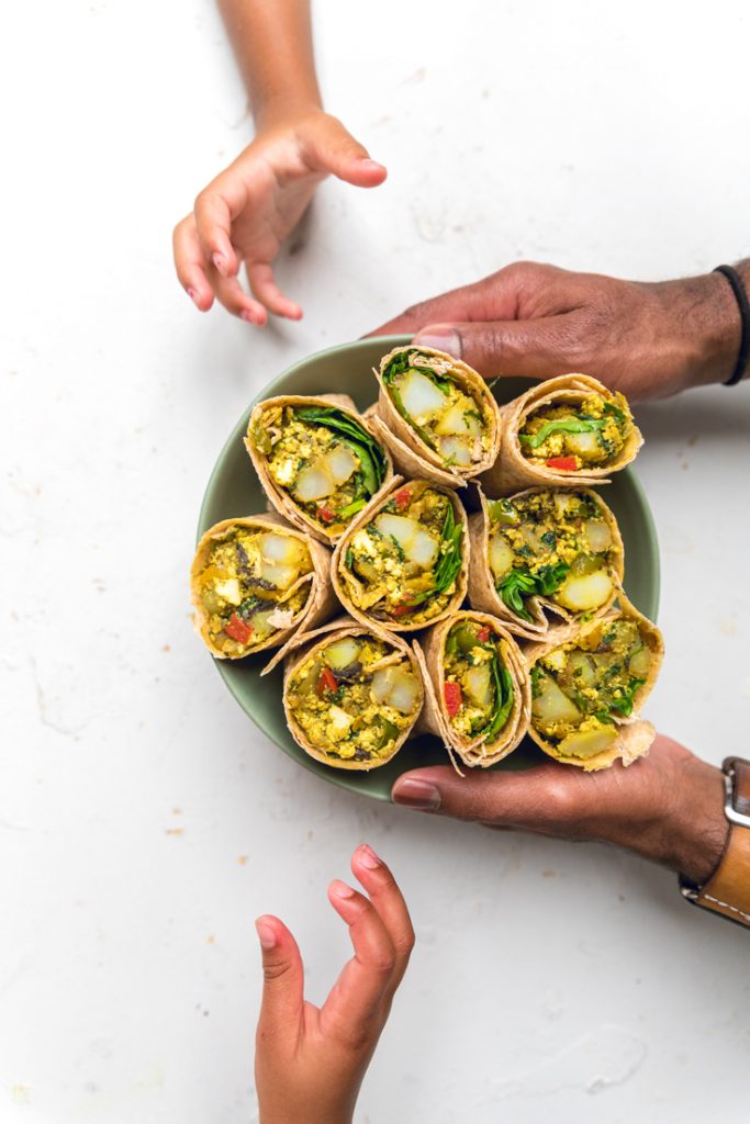 Hands holding a bowl of vegan tofu breakfast burritos with two kids hands reaching for bowl.