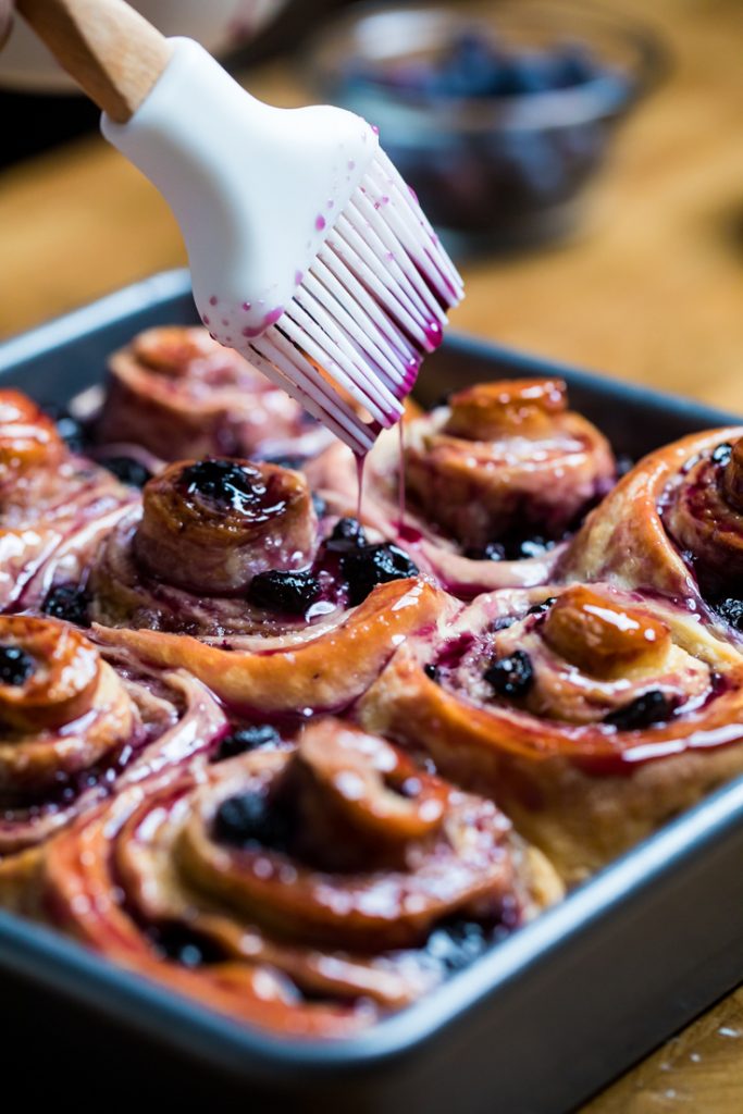 A pastry brush brushing on simple syrup of vegan blueberry cinnamon rolls.
