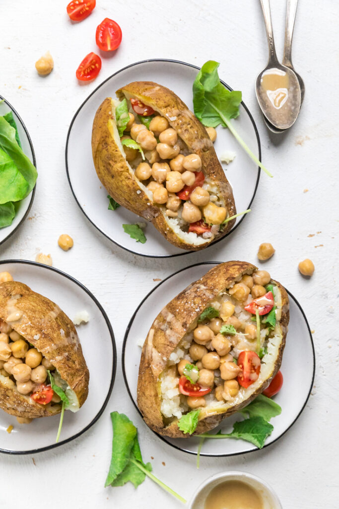 Vegan stuffed baked potatoes on white plates topped with chickpeas, tomatoes, spinach and tahini.