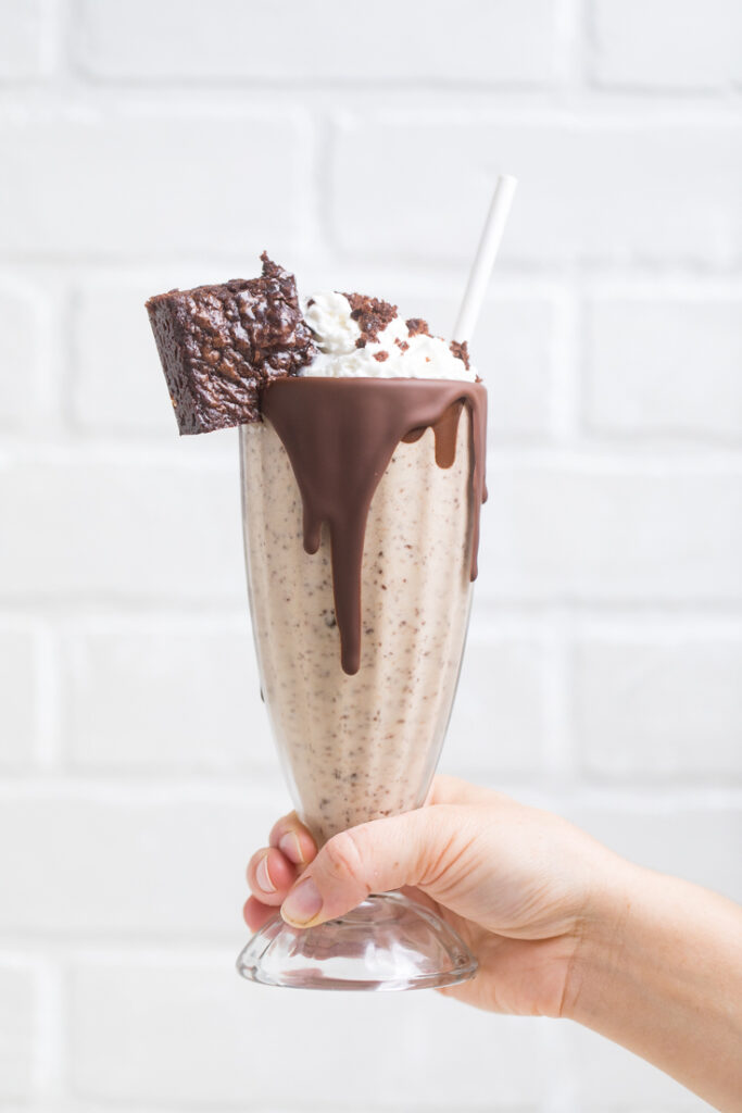 A hand holding a vegan peanut butter fudge brownie milkshake topped with vegan brownie on side.