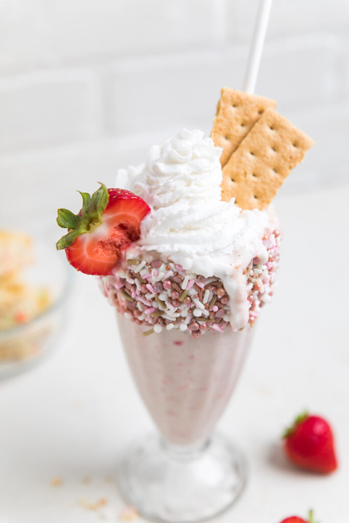 A Vegan strawberry birthday cake milkshake topped with vegan whipped cream, fresh strawberries and graham crackers.