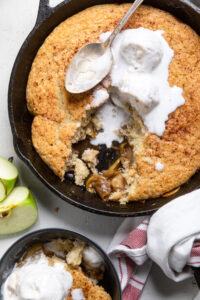 Vegan apple cobbler in a skillet with vanilla ice cream and a piece missing.