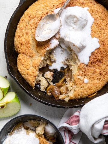 Vegan apple cobbler in a skillet with vanilla ice cream and a piece missing.