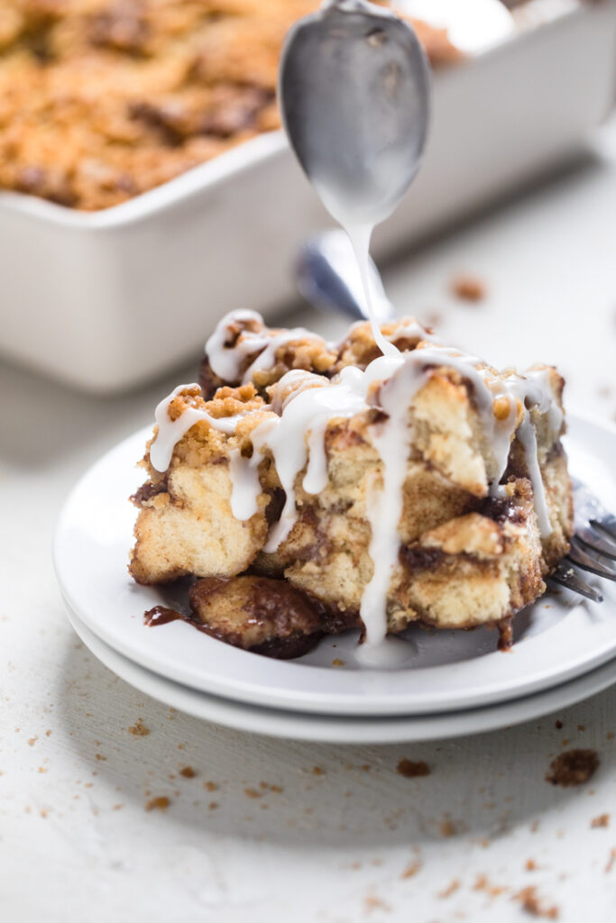A serving of vegan cinnamon roll casserole with a spoon drizzling glaze over top.