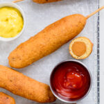 A batch of vegan corn dogs with bowls of ketchup and mustard.