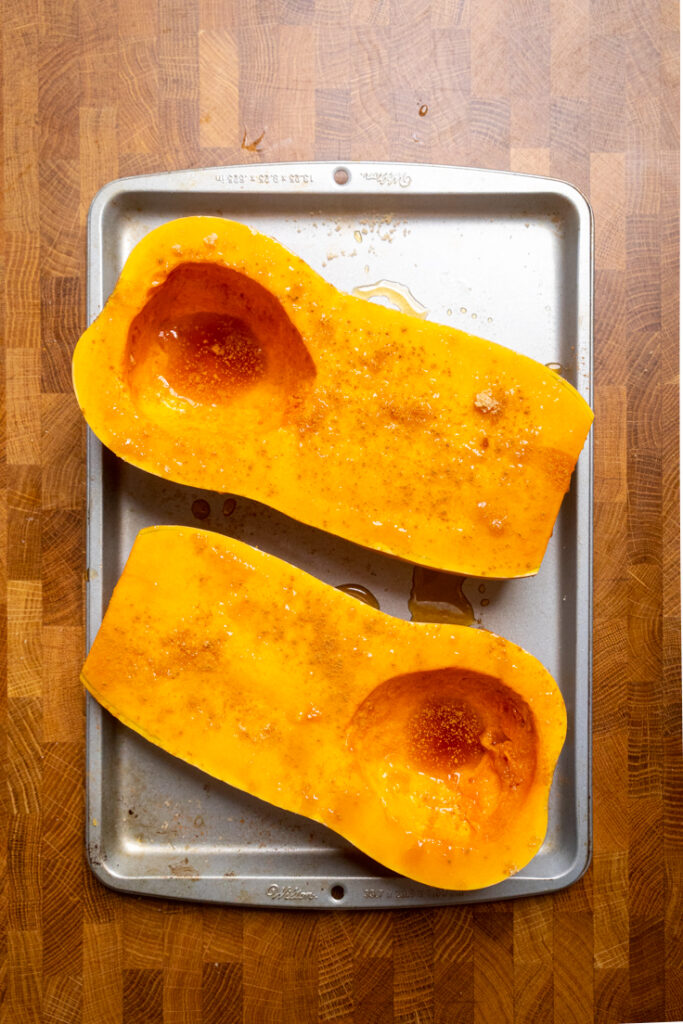 Uncooked Butternut Squash halved on a tray with maple syrup salt and pepper.