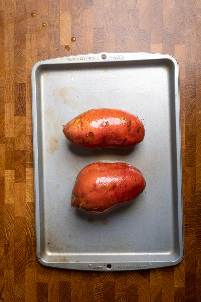 Two sweet potatoes on a tray with oil brushed.