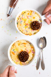 Two servings of vegan vanilla mug cake with rainbow sprinkles and chocolate frosting on top.