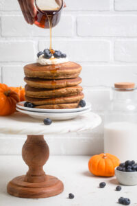 A stack of Vegan Pumpkin Pancakes with frosting, blueberries and syrup.