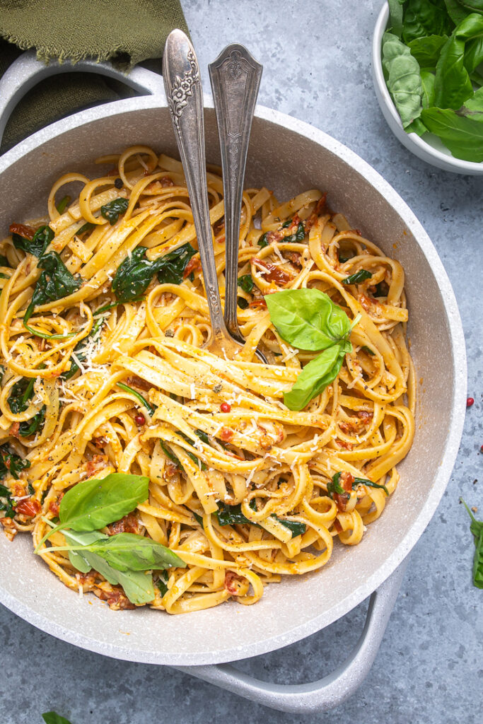 A gray pot of vegan spinach and sundried tomato pasta with two spoons inside. 
