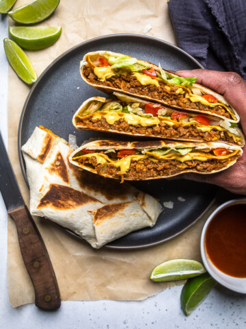A hand holding three pieces of Vegan crunchwrap supremes over a plate.