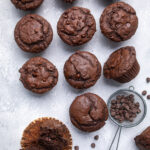 Seven vegan double chocolate muffins on a table with a sieve of chocolate chips.