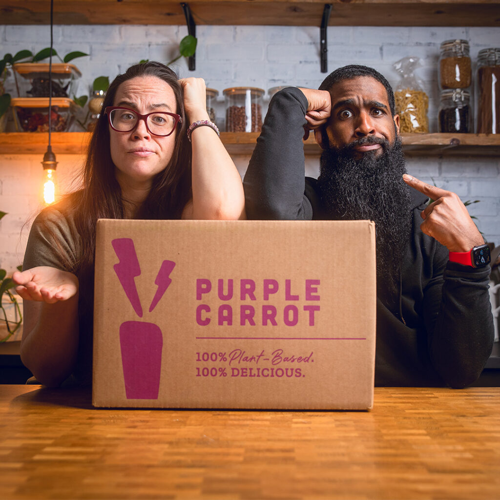 A couple standing behind a box of purple carrot meals making funny faces.