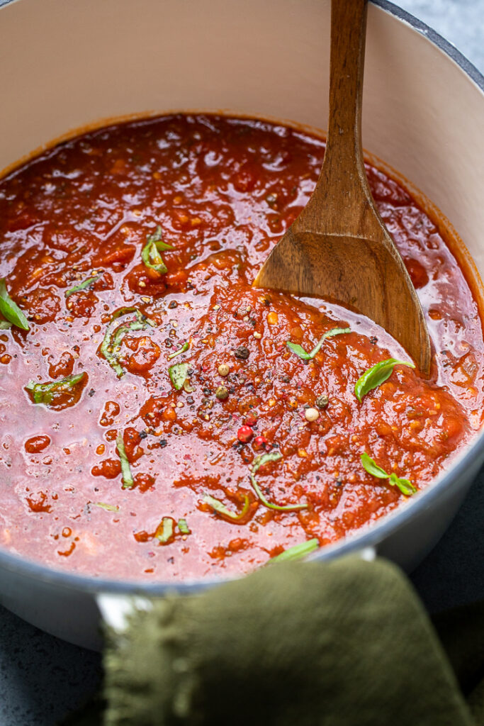 A wooden spoon inside a pot of spaghetti sauce with fresh basil and black peppercorns.
