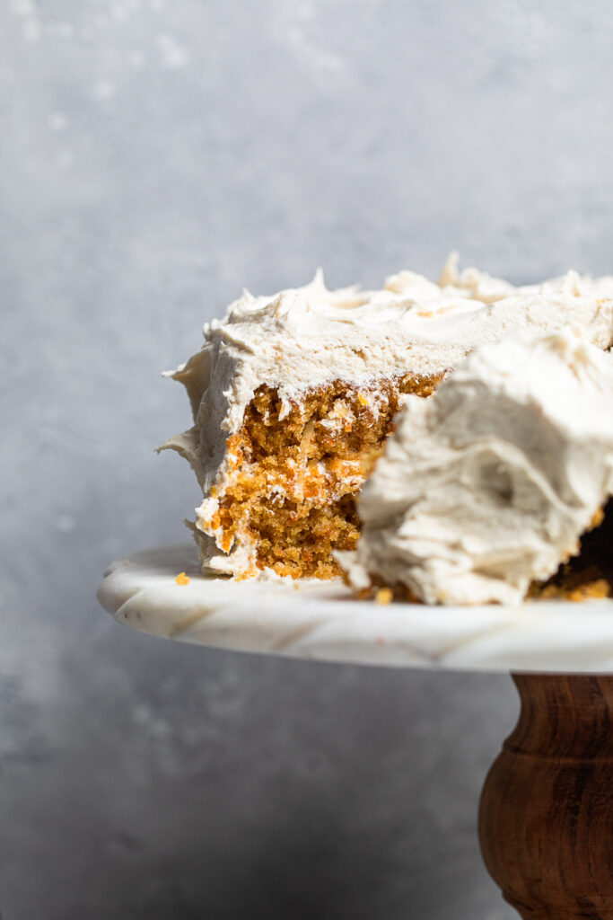 A vegan coconut carrot cake on a white cake stand with a slice missing.