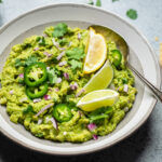 A gray and white bowl of copycat chipotle guacamole with slices lime and lemon wedges.
