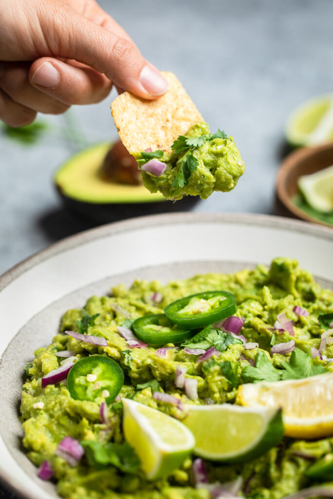 A hand holding a chip with copycat chipotle guacamole over a bowl.