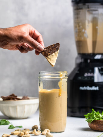 A tortilla chip being held over a jar of vegan cashew queso.