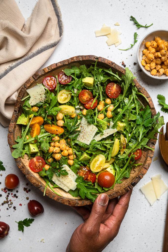 A bowl of vegan arugula salad with freshly sliced vegan parmesan, chickpeas, tomatoes and avocado.