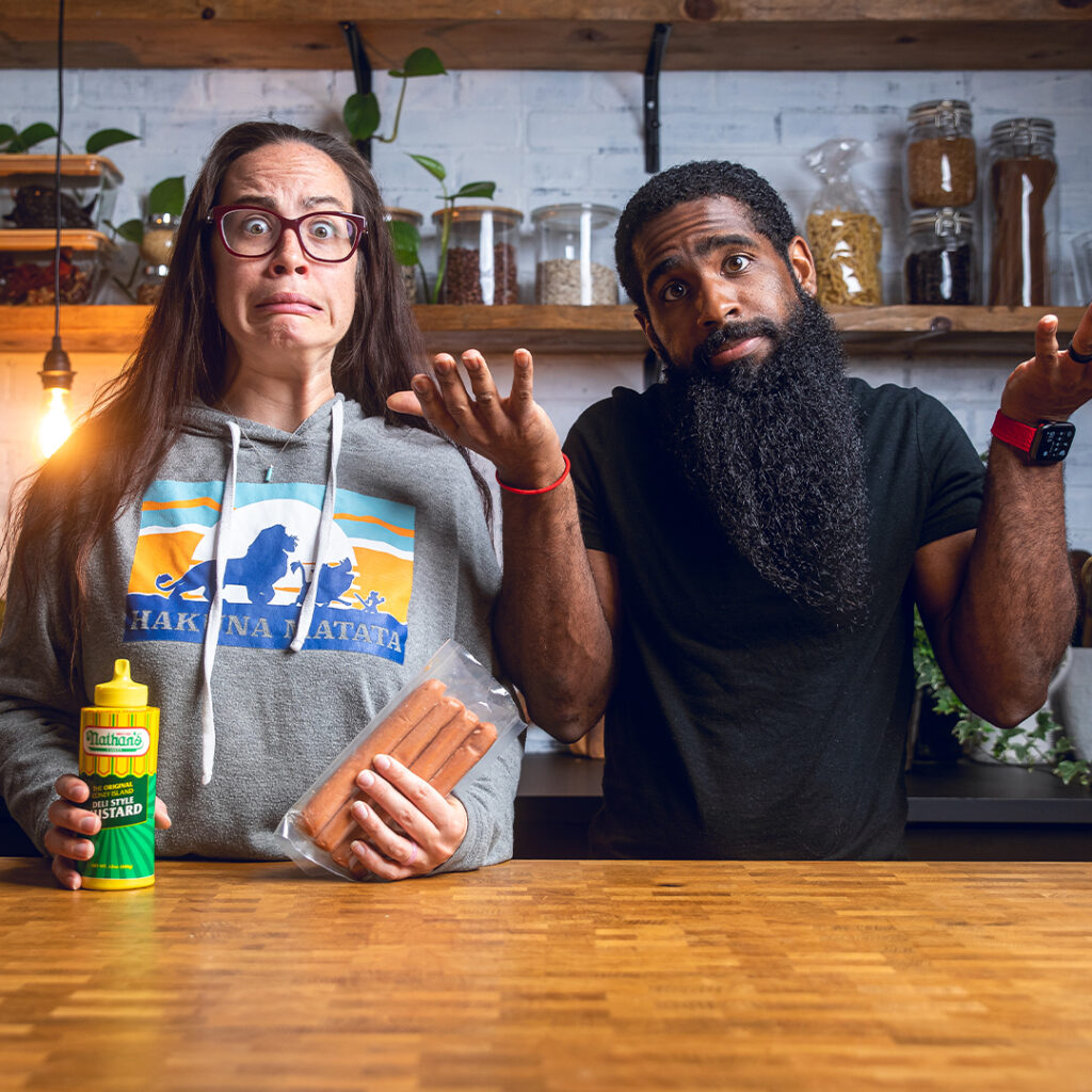 A couple standing behind a table making silly faces while holding vegan hot dogs and mustard.
