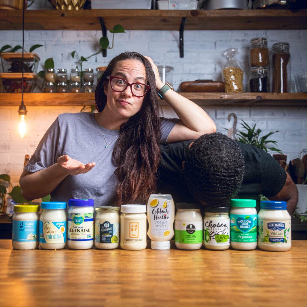A couple posing behind a table of vegan mayo bottles making silly faces. 