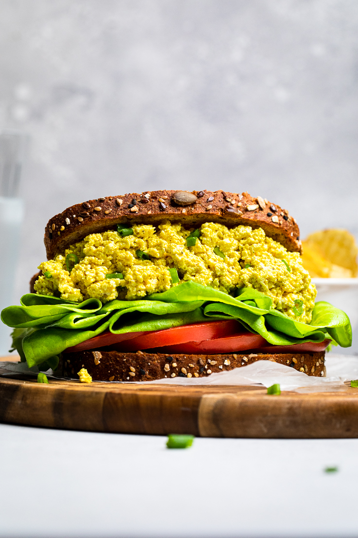 A flat easy vegan egg salad sandwich with lettuce and tomato on a board.