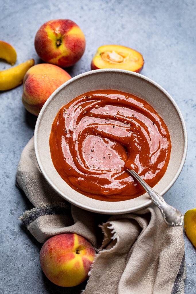 A gray bowl of vegan peach BBQ sauce with fresh peaches on the table around.