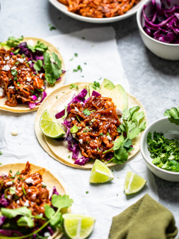 Three grilled vegan jackfruit tacos laying flat on a table with fresh cilantro and limes.