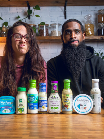 A couple making funny faces sitting behind a table of vegan ranch dressing product packages.