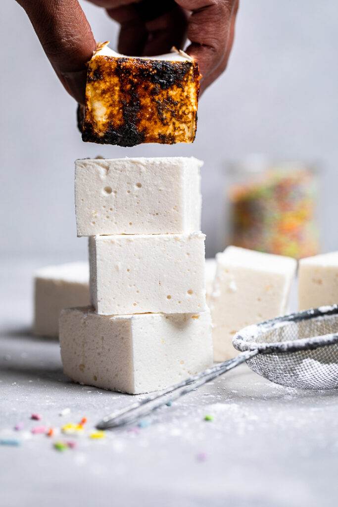 A hand placing a charred vegan marshmallow to place on a stack of uncharred marshallows.