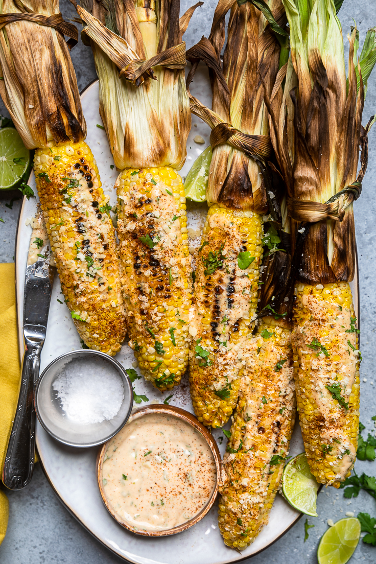 A tray of vegan grilled Mexican street corn with husks intact and charred and dipping sauces.