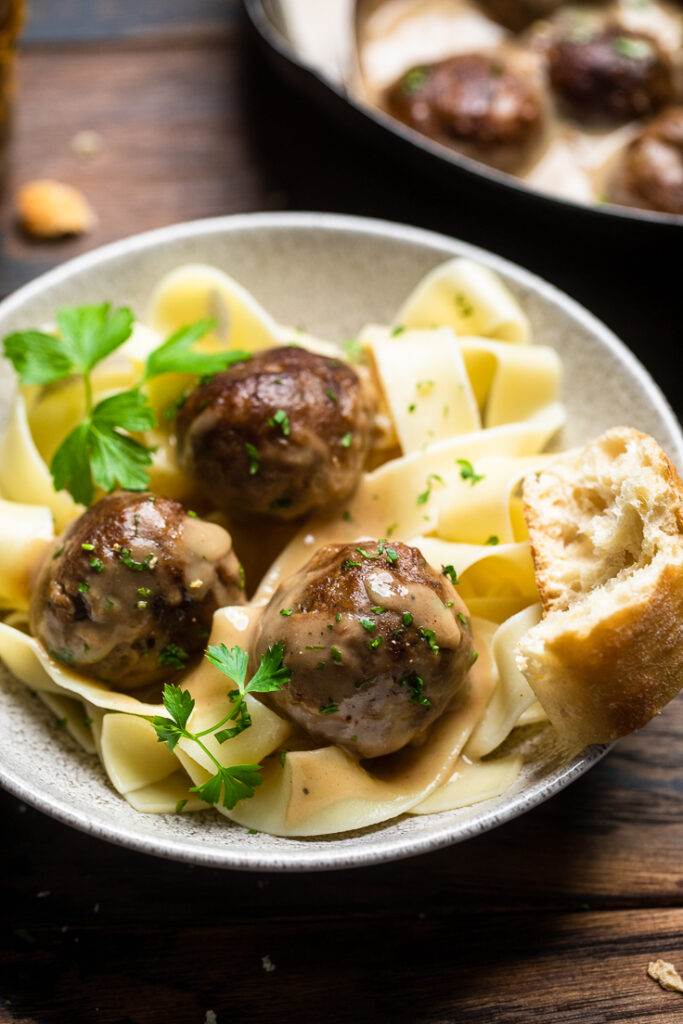 A bowl of saucy vegan Swedish meatballs over vegan pasta and a piece of crunchy bread.
