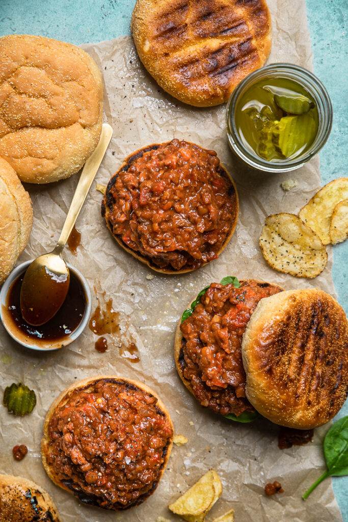 Arrangement of slow cooker vegan lentil sloppy joe sandwiches on a table with chips and pickles.